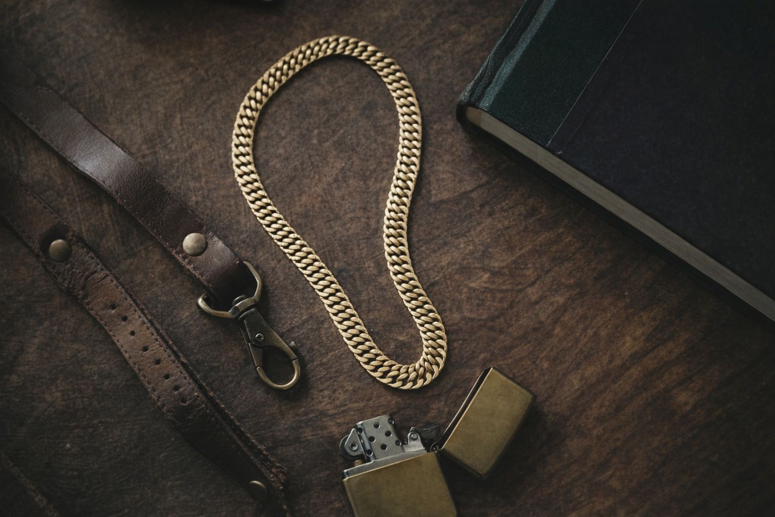 Guldpläterat bismarckhalsband på rustik träyta bredvid en tändare och läderrem.